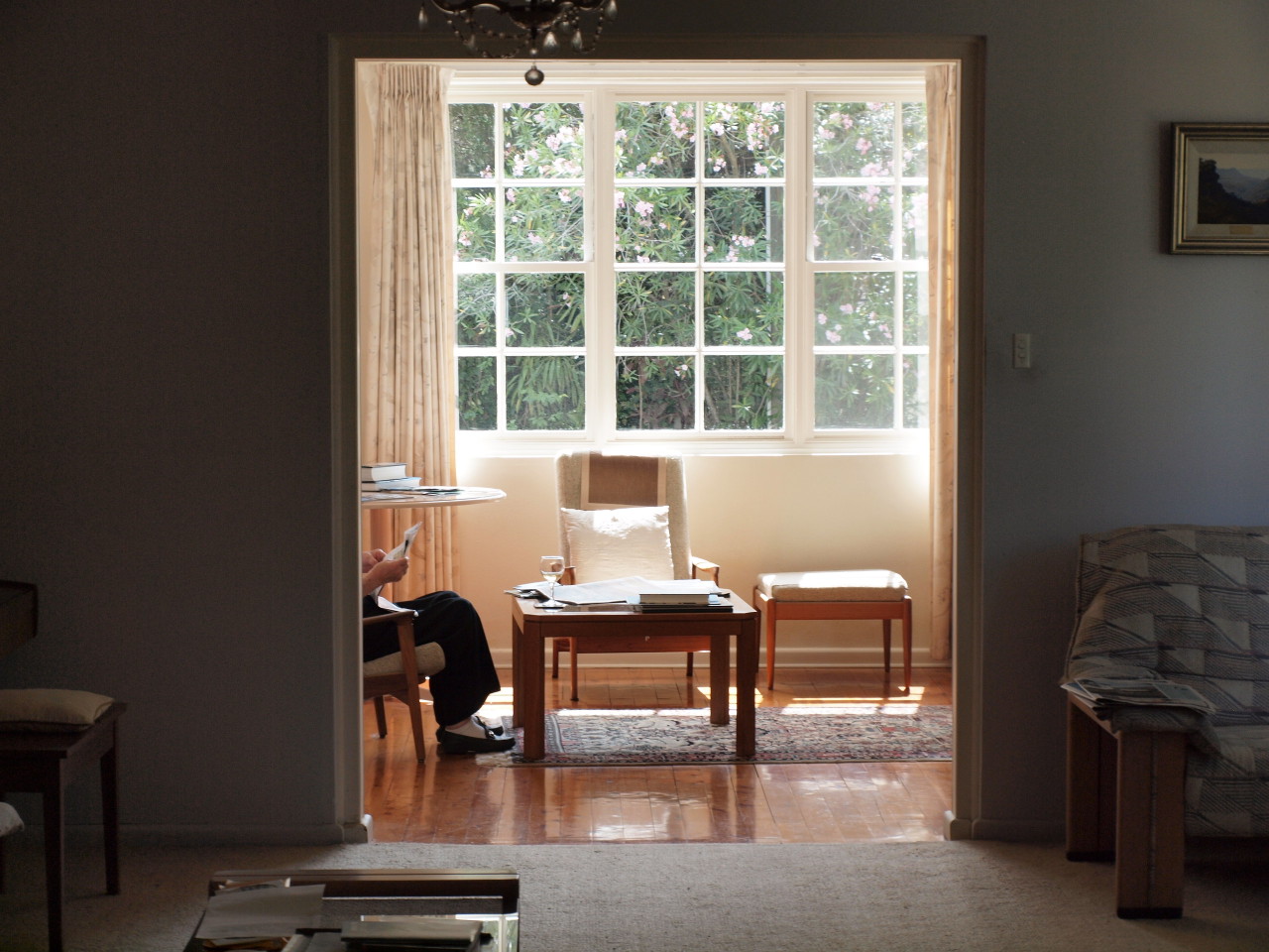 Interior home shot: an adult sits in a chair framed by a doorway, with a large window providing natural light and a view to greenery.