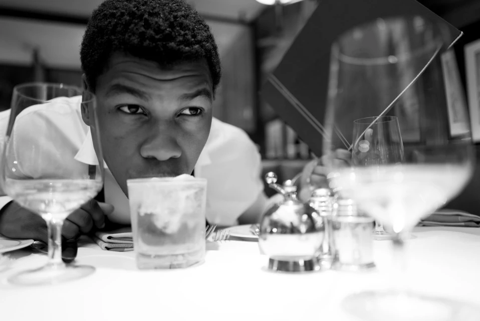 Black and white image of a man in a restaurant setting with wine glasses, a cocktail, and other tableware on a white tablecloth.
