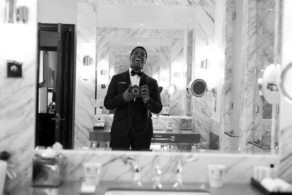 Smiling man in a tuxedo takes a mirror selfie in a marble bathroom. He's holding a camera, looking at his reflection with an open-mouth smile.