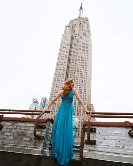 A woman in a blue gown poses on a rooftop with the Empire State Building in the background on a cloudy day in an eye-level full shot.