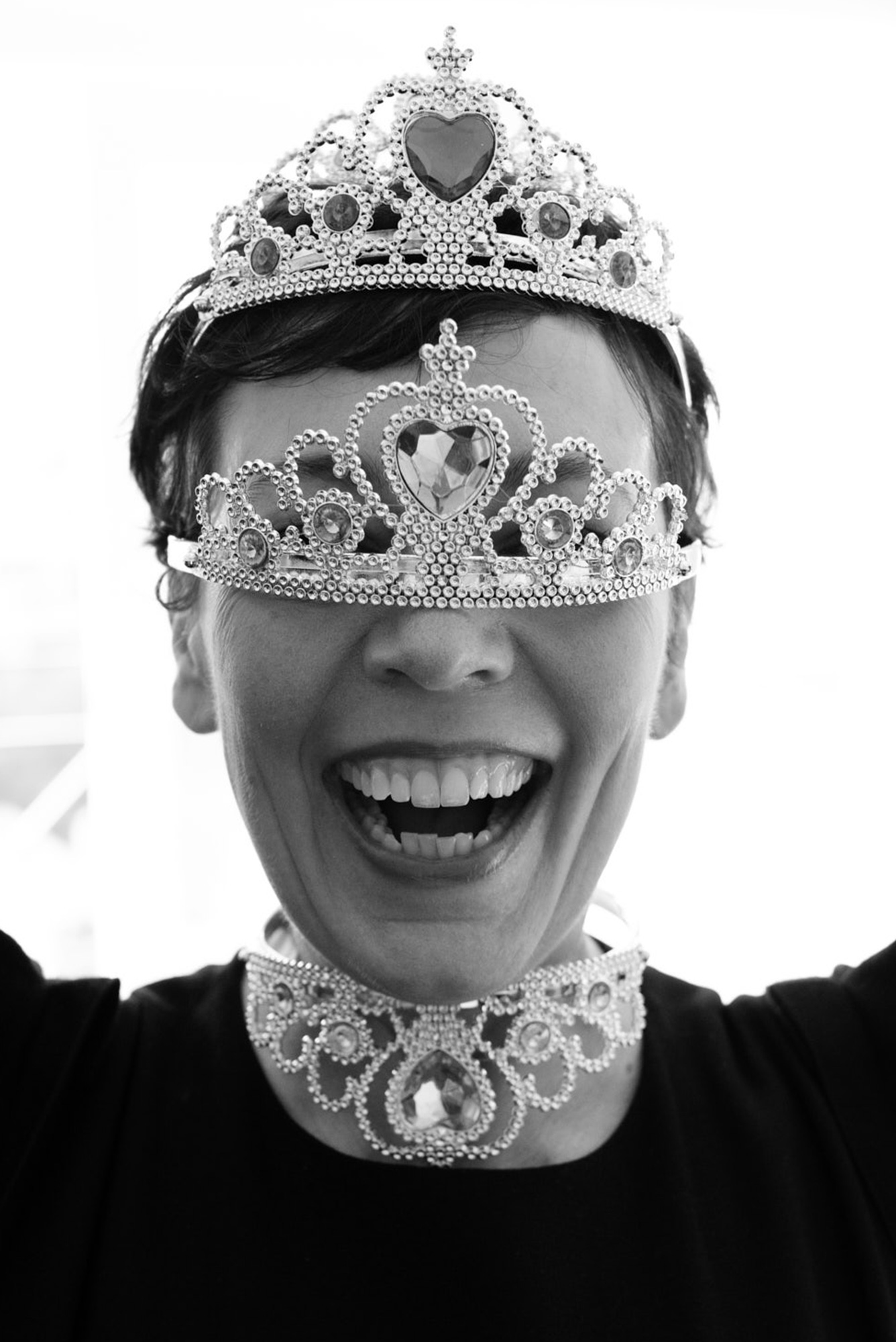Woman with open mouth wearing a tiara, choker, and mask. Black and white photo of a person with fashion accessories and dark clothing.