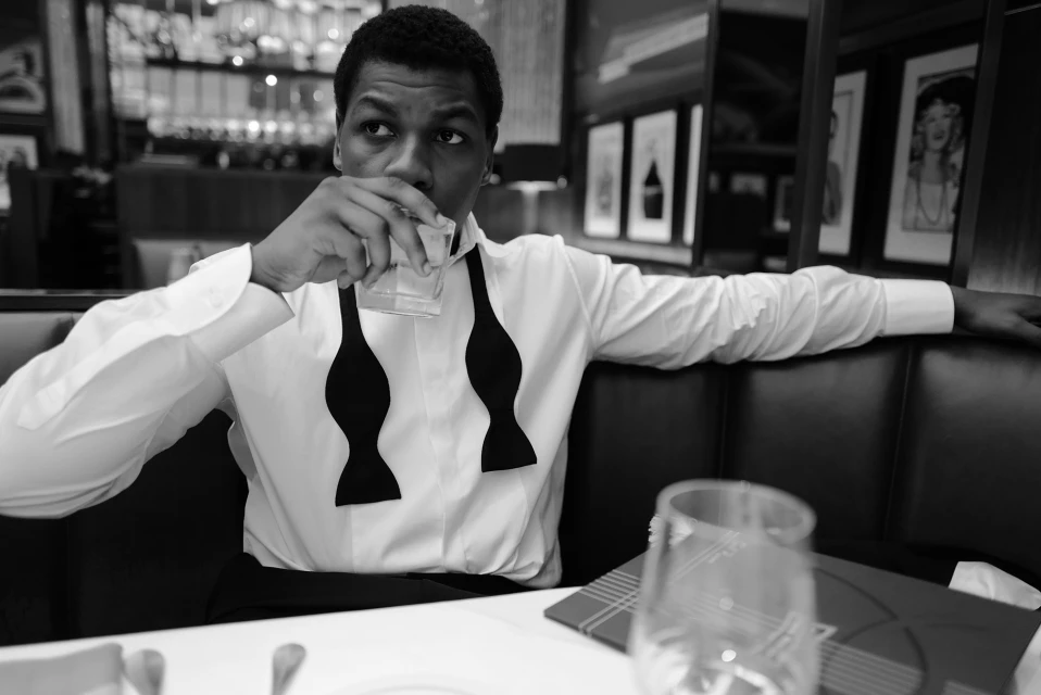 Monochrome portrait of a Black adult male in a white dress shirt and black bow ties, seated indoors while drinking from a glass.