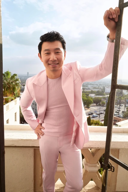 Headshot of Asian male in pink suit on outdoor balcony with city skyline backdrop and clear sky, engaging with a gentle smile.