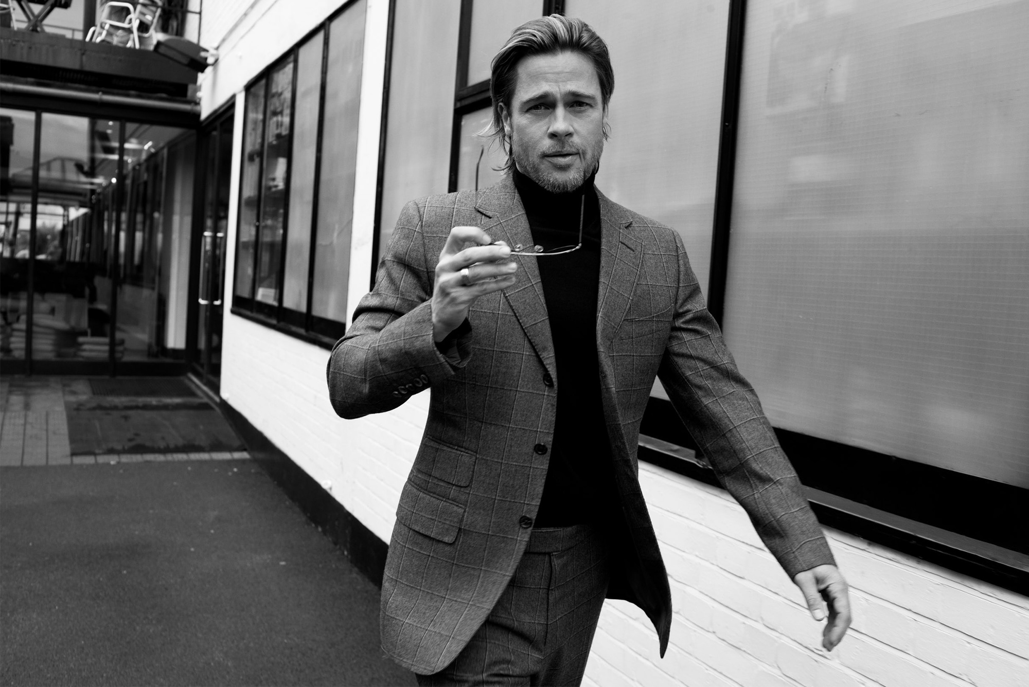 Black and white portrait of a mature adult male wearing a suit, holding glasses, and walking in an urban environment.