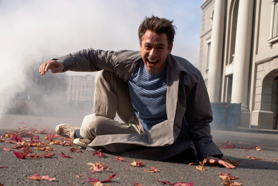 An adult male in a grey trench coat and tan pants is sitting on the ground laughing with fall leaves scattered around him and smoke in background.