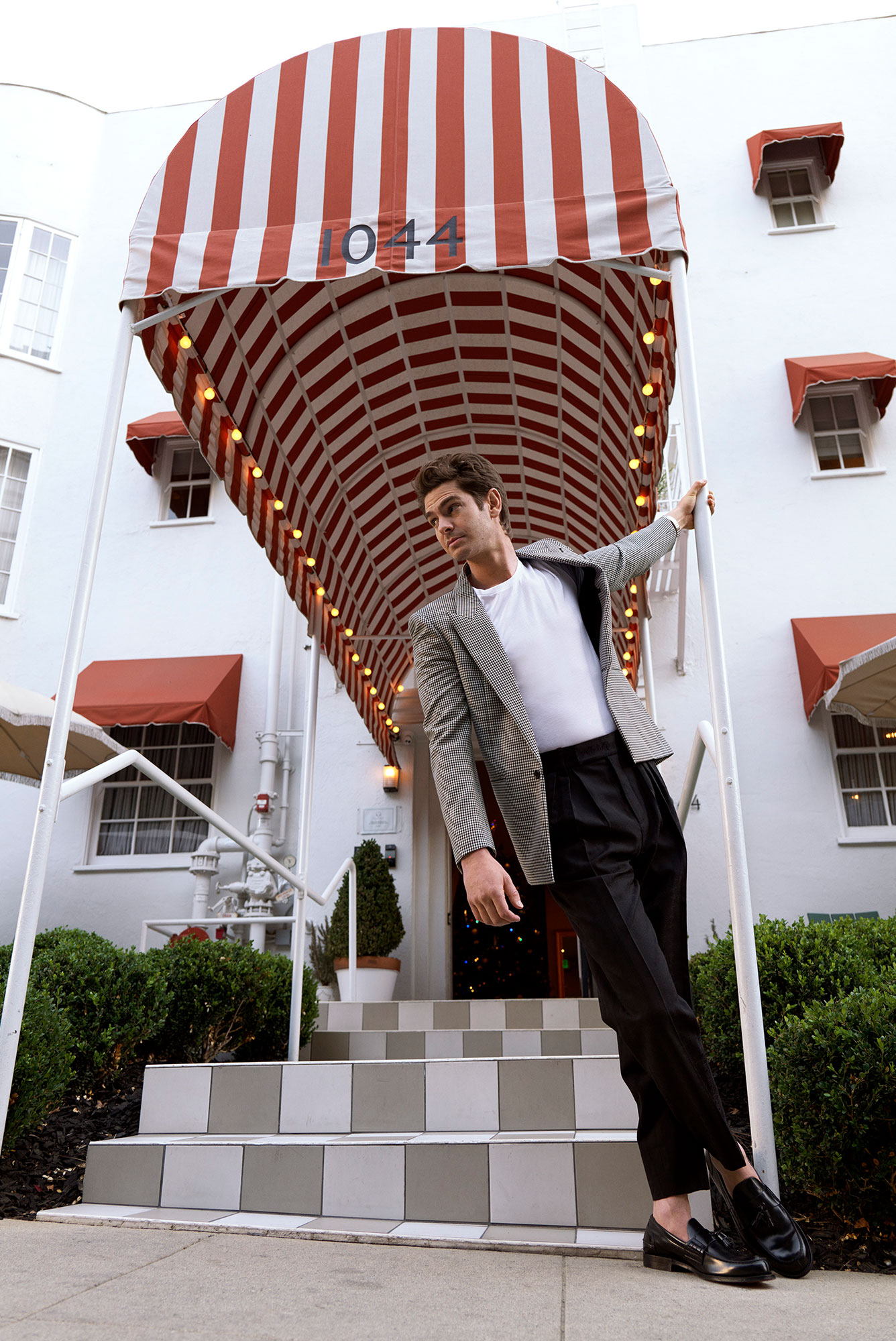 Man in checkered blazer poses in front of white building with striped awning and gray tiled steps. Stylish fashion and architecture.
