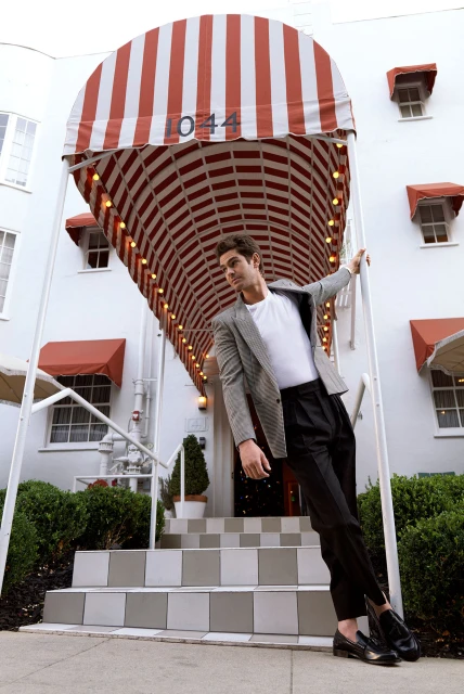 Man in checkered blazer poses in front of white building with striped awning and gray tiled steps. Stylish fashion and architecture.
