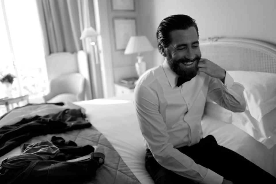 Black-and-white shot of a smiling man with a beard sitting on a bed in a dress shirt, adjusting his bow tie.