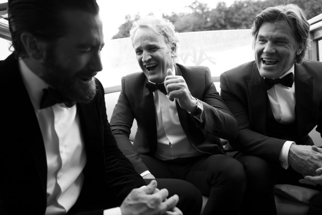 Three adult men in tuxedos laugh together while seated. The black and white photo captures a light moment between the men.