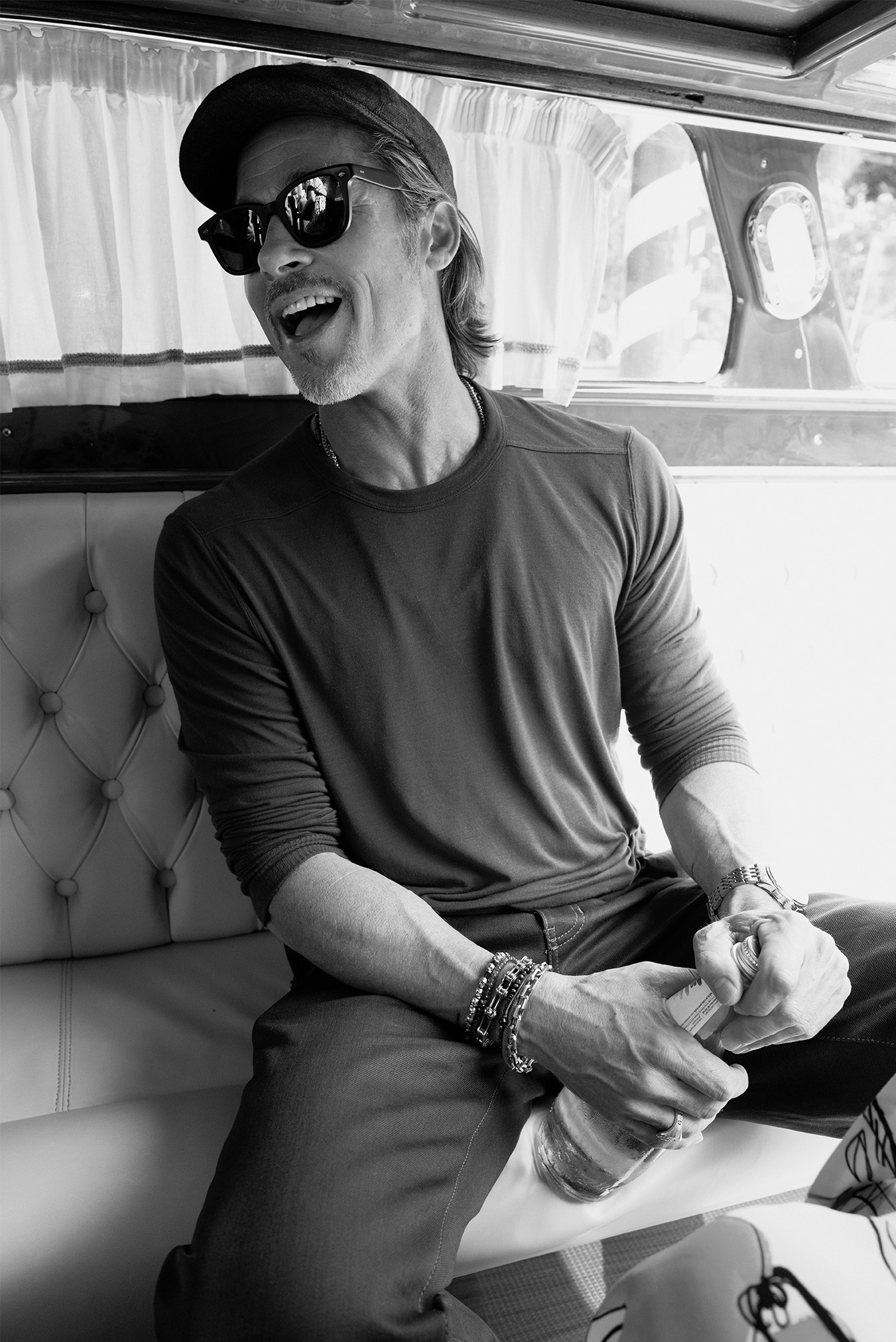 Monochromatic shot of a man wearing sunglasses and a baseball cap, sitting in a boat and smiling while holding a bottle.