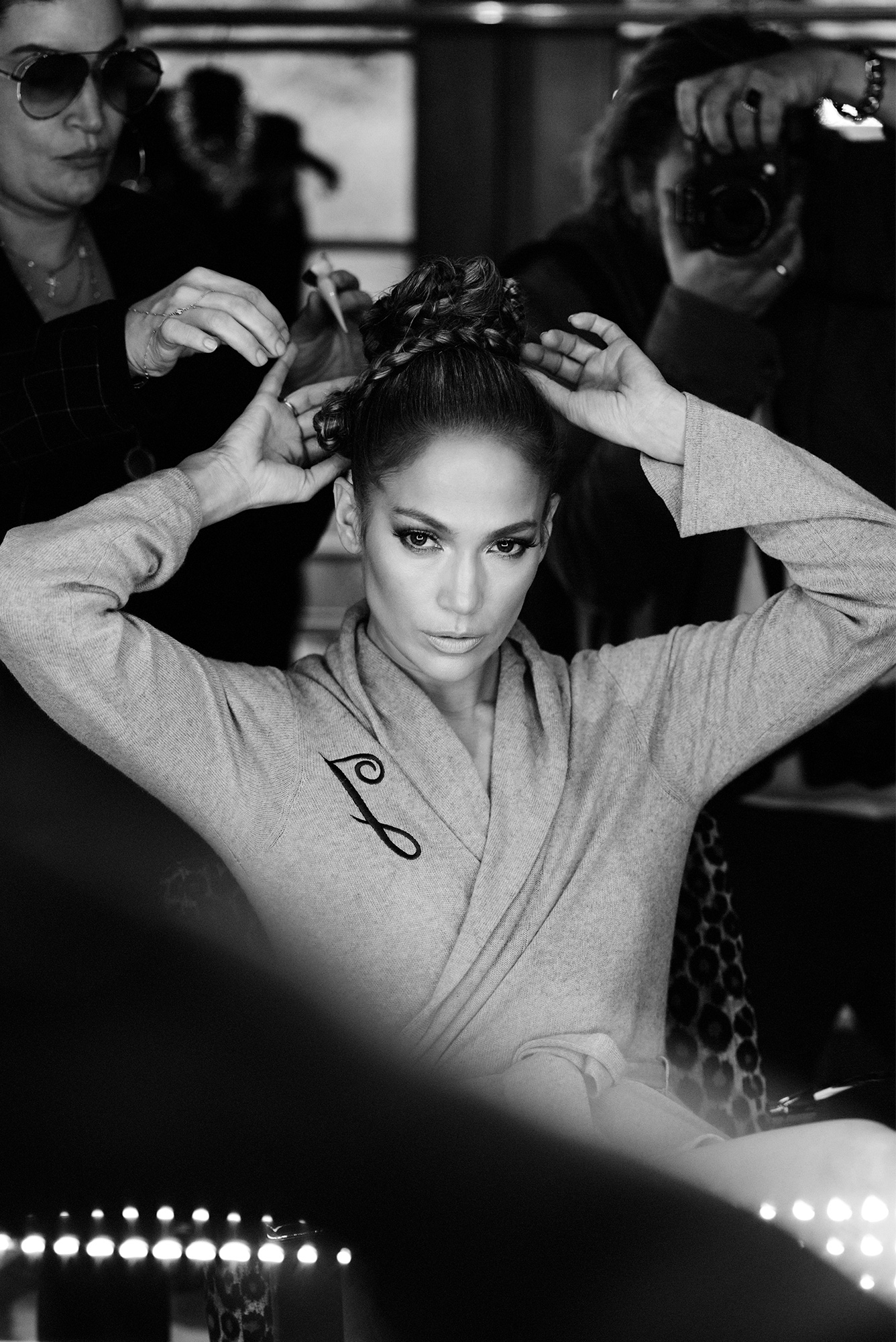 Black and white portrait of Jennifer Lopez getting her hair done. She is wearing a gray robe with a black “J” on it.