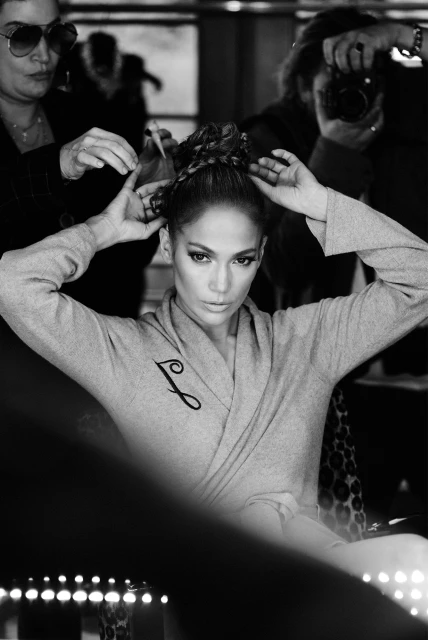 Black and white portrait of Jennifer Lopez getting her hair done. She is wearing a gray robe with a black “J” on it.
