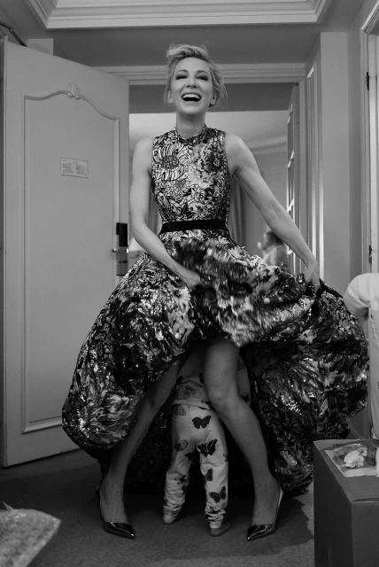Black and white shot of a smiling woman in a formal dress playfully holding the dress up to reveal a baby underneath it.