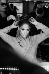 Monochrome backstage portrait of Jennifer Lopez styling her hair with crew members visible in the background
