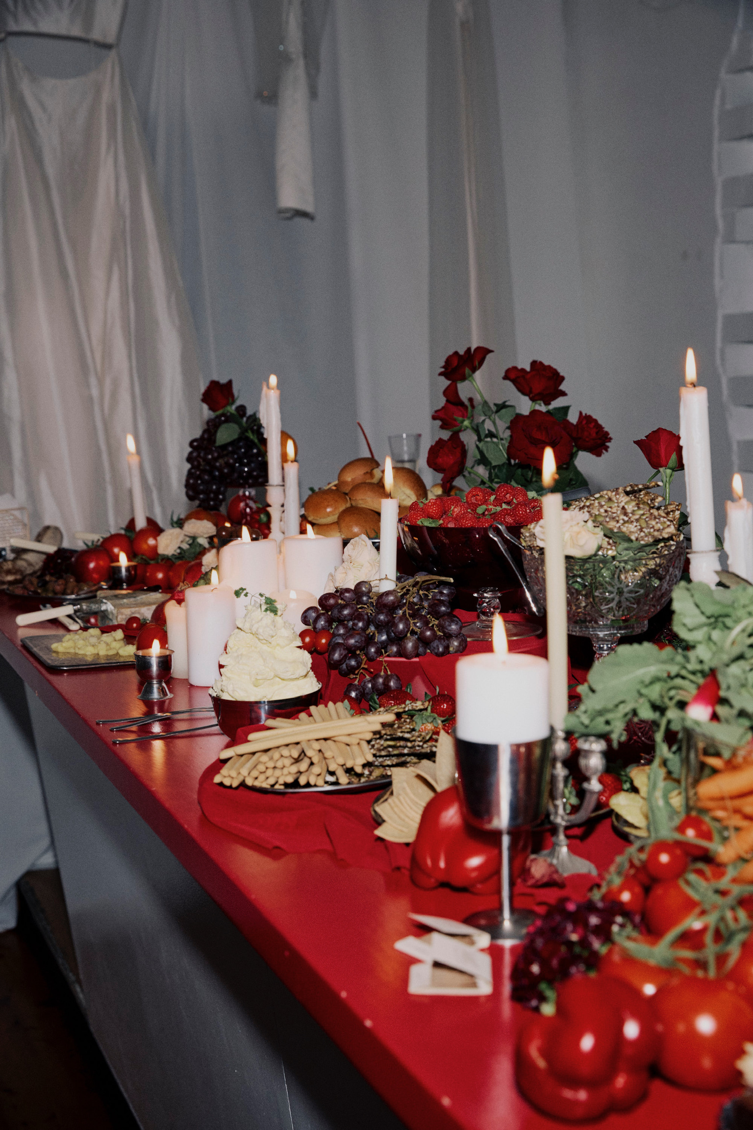 Table setting with candles, roses, grapes, crackers, tomatoes, and cheese on a red table in front of a white curtain backdrop.