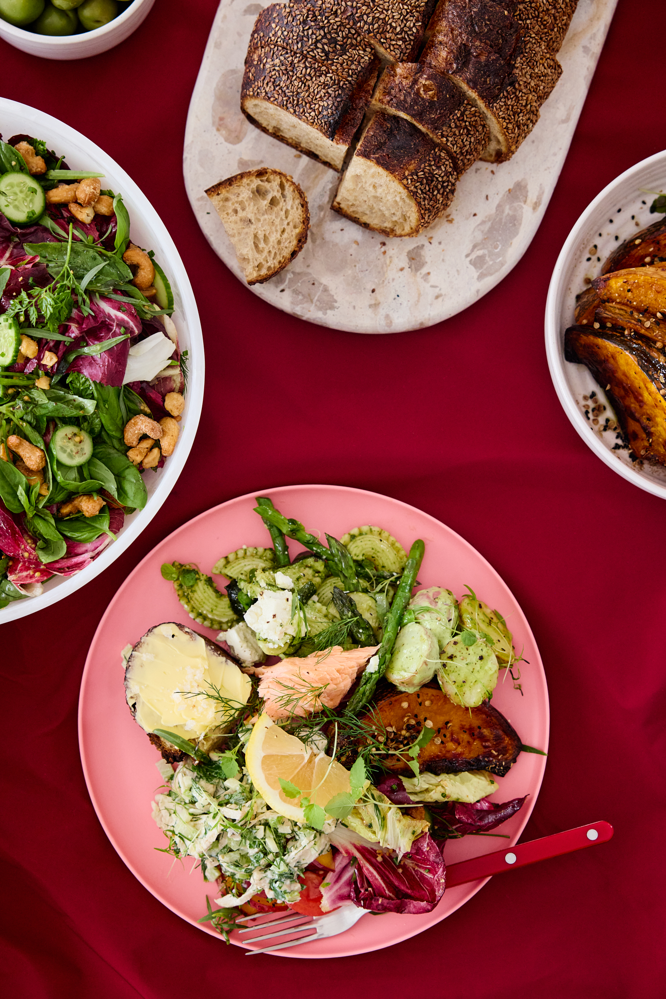 Overhead shot of a pink plate with salmon, potato, and mixed greens on a red tablecloth. Bowls of salad and sliced bread visible.