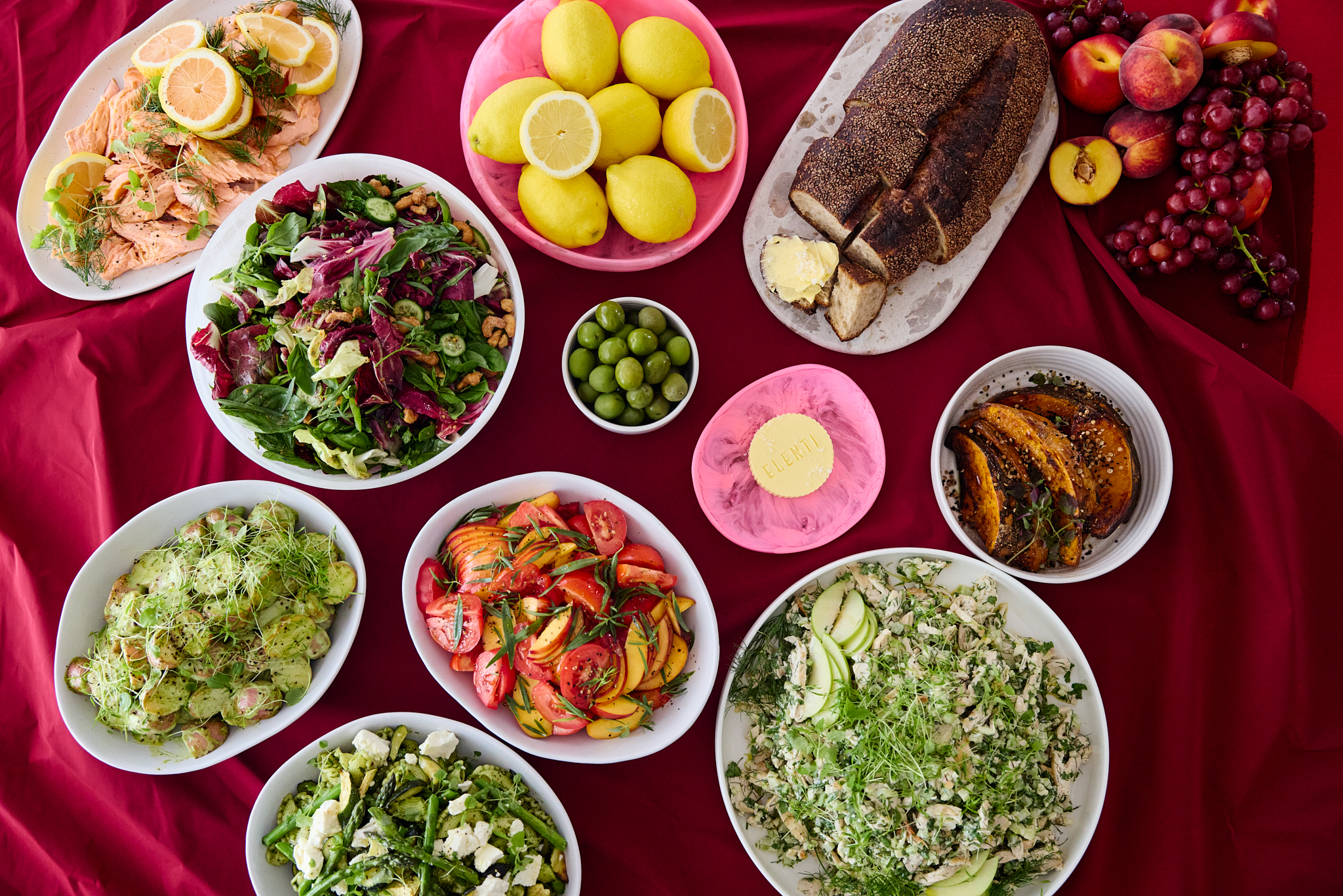 Overhead view of an array of fresh food bowls and platters arranged on a red tablecloth, including salad, salmon, lemons, and bread.