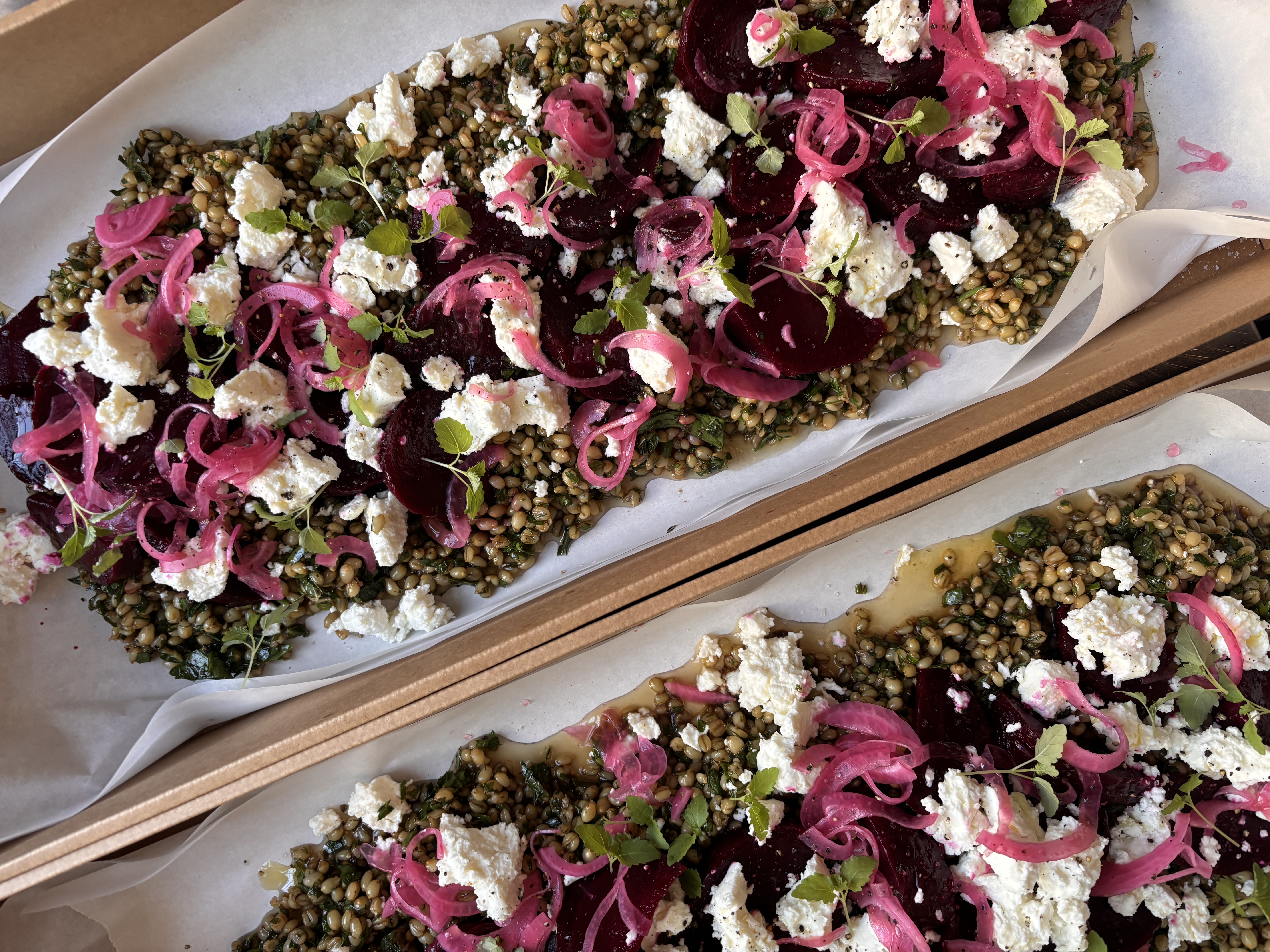 Two food trays hold flatbreads with beet slices, pickled onions, herbs, and crumbled cheese, presented in a restaurant or catering setting.