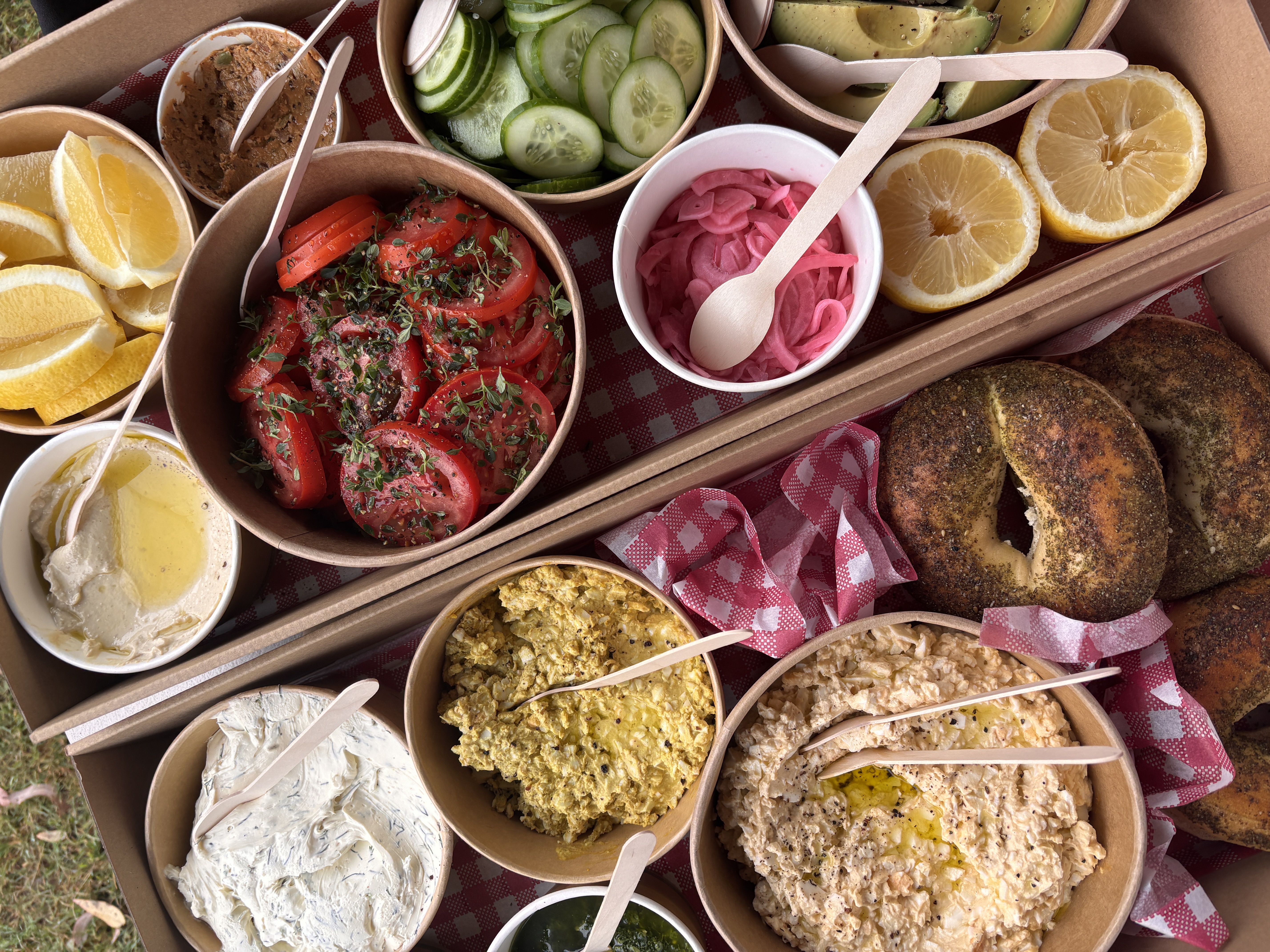 Overhead shot of a picnic box filled with an assortment of dips, sliced vegetables, lemons, and bagels, perfect for a fresh, outdoor meal.