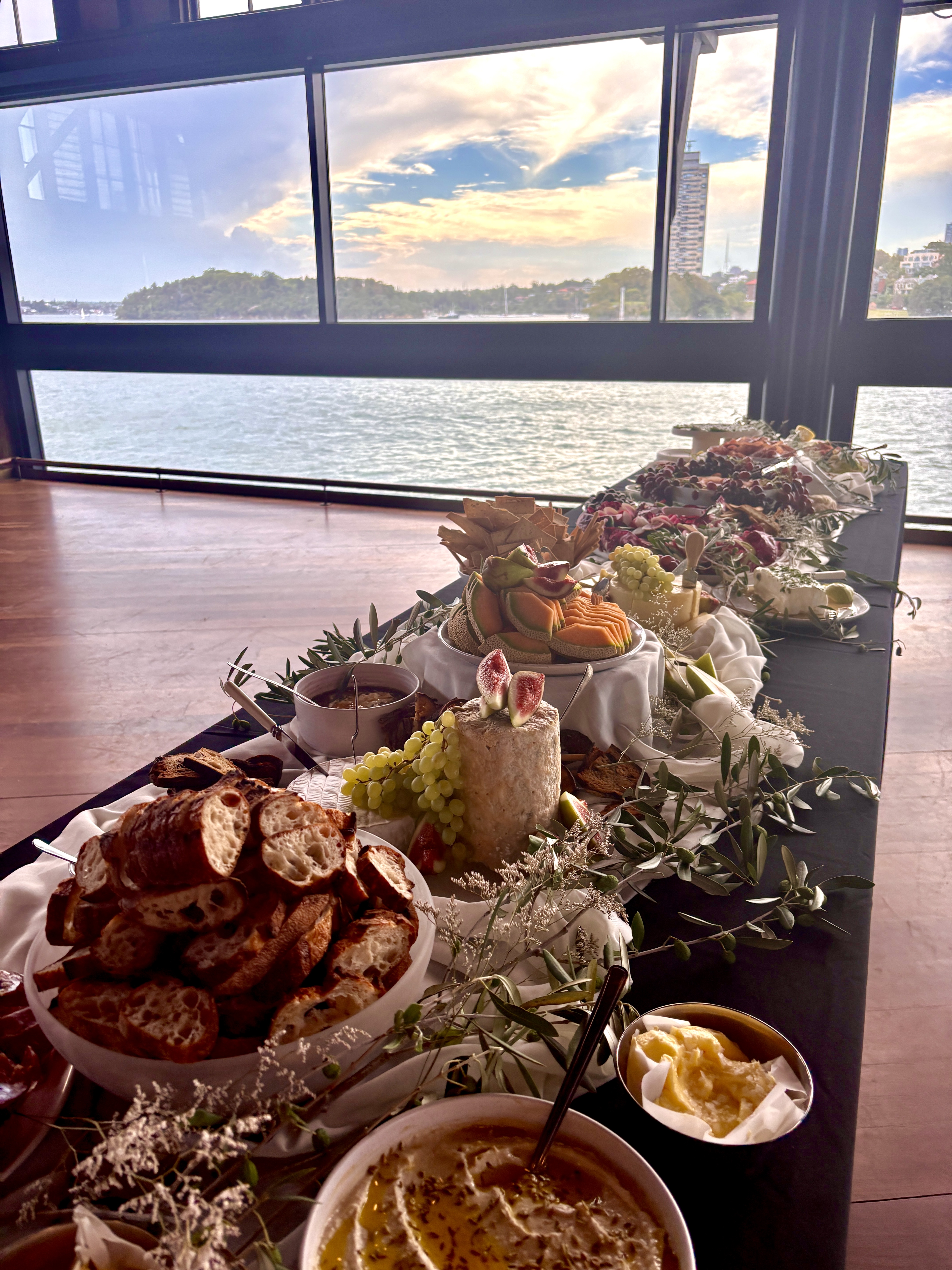 Indoor catering setup with table filled with bread, hummus, cheese, and fruit, set against a window view of the ocean and cityscape.