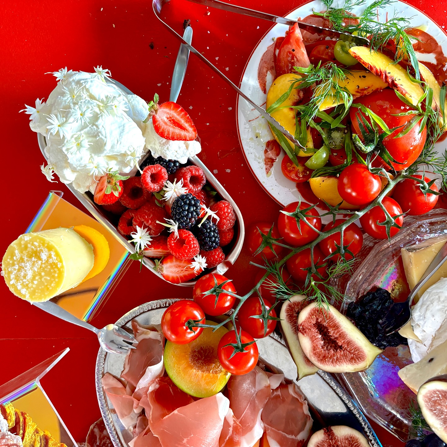 Overhead shot of an assortment of charcuterie, cheeses, and fresh fruits, including figs, berries, and melon, arranged on a red table.