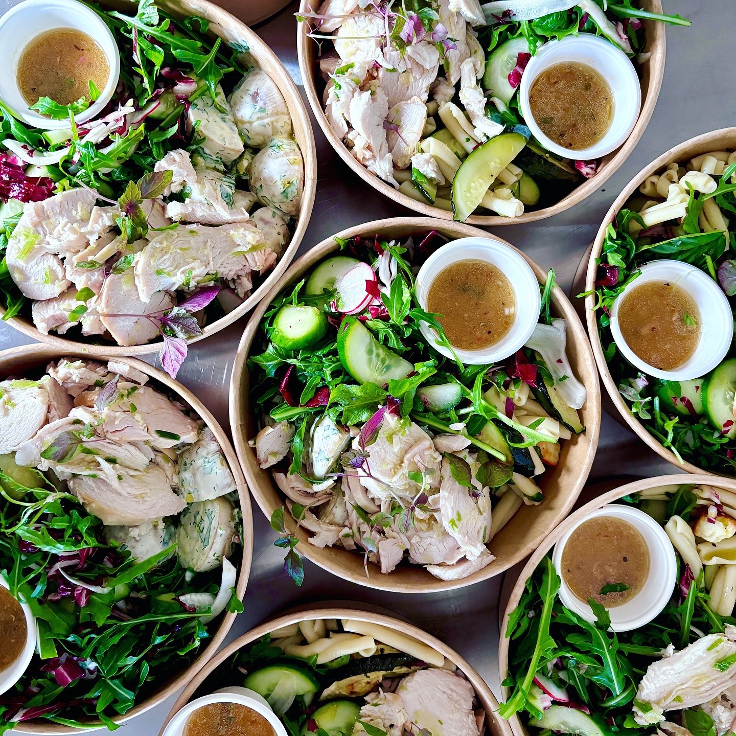 Overhead shot of multiple salad bowls with mixed greens, grains, sliced chicken, cucumber, and a small sauce container in each.