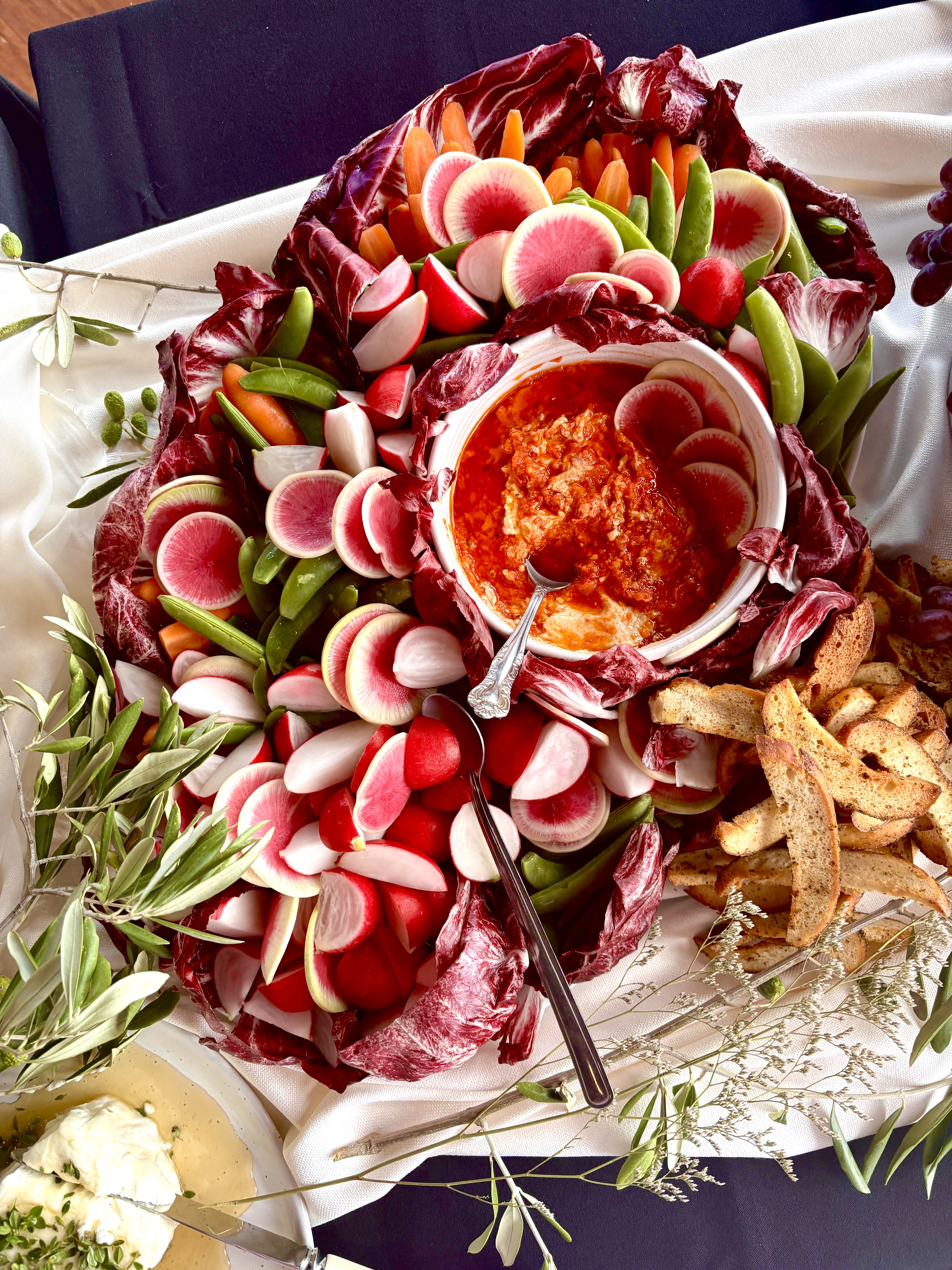 Overhead shot of an appetizer platter with radish, carrot sticks, snow peas, crackers and dip arranged on a white tablecloth.