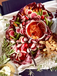 An overhead food spread featuring sliced watermelon radishes, baby carrots, snap peas, bread crisps, and a central dip bowl.