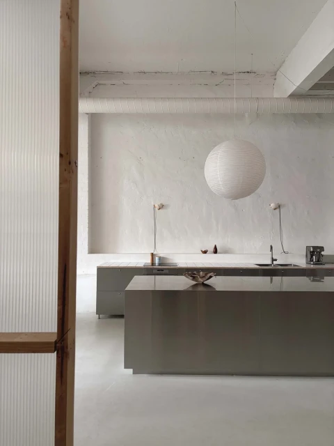 Minimalist kitchen interior featuring gray cabinets, white walls, hanging paper lantern, and wooden door frame in natural light.