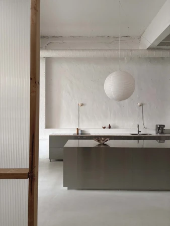 Minimalist kitchen interior featuring gray cabinets, white walls, hanging paper lantern, and wooden door frame in natural light.
