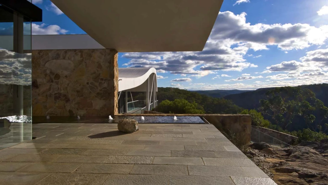 Exterior view of a modern architectural design featuring a stone terrace with a small fountain, overlooking a lush mountainous landscape.