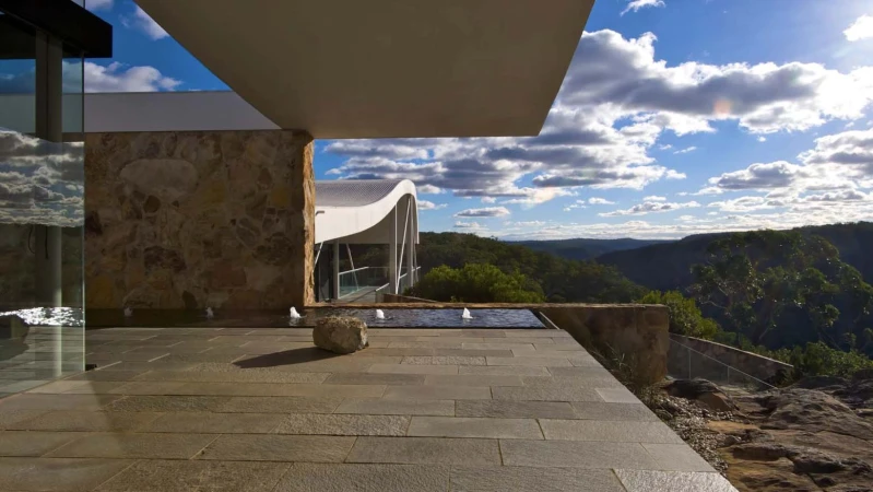 Exterior view of a modern architectural design featuring a stone terrace with a small fountain, overlooking a lush mountainous landscape.