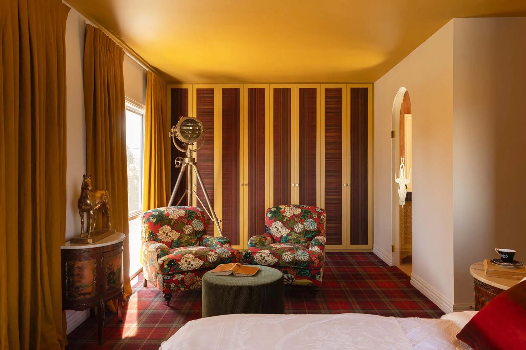 Bedroom interior with floral armchairs, a floor lamp, a patterned carpet, and a window with golden drapes, creating a cozy atmosphere.