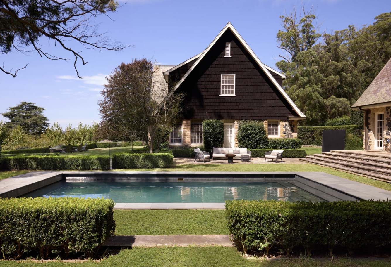 Exterior shot of a two-story house with dark brown shingles, a stone facade, and a pool in the foreground on a sunny day.