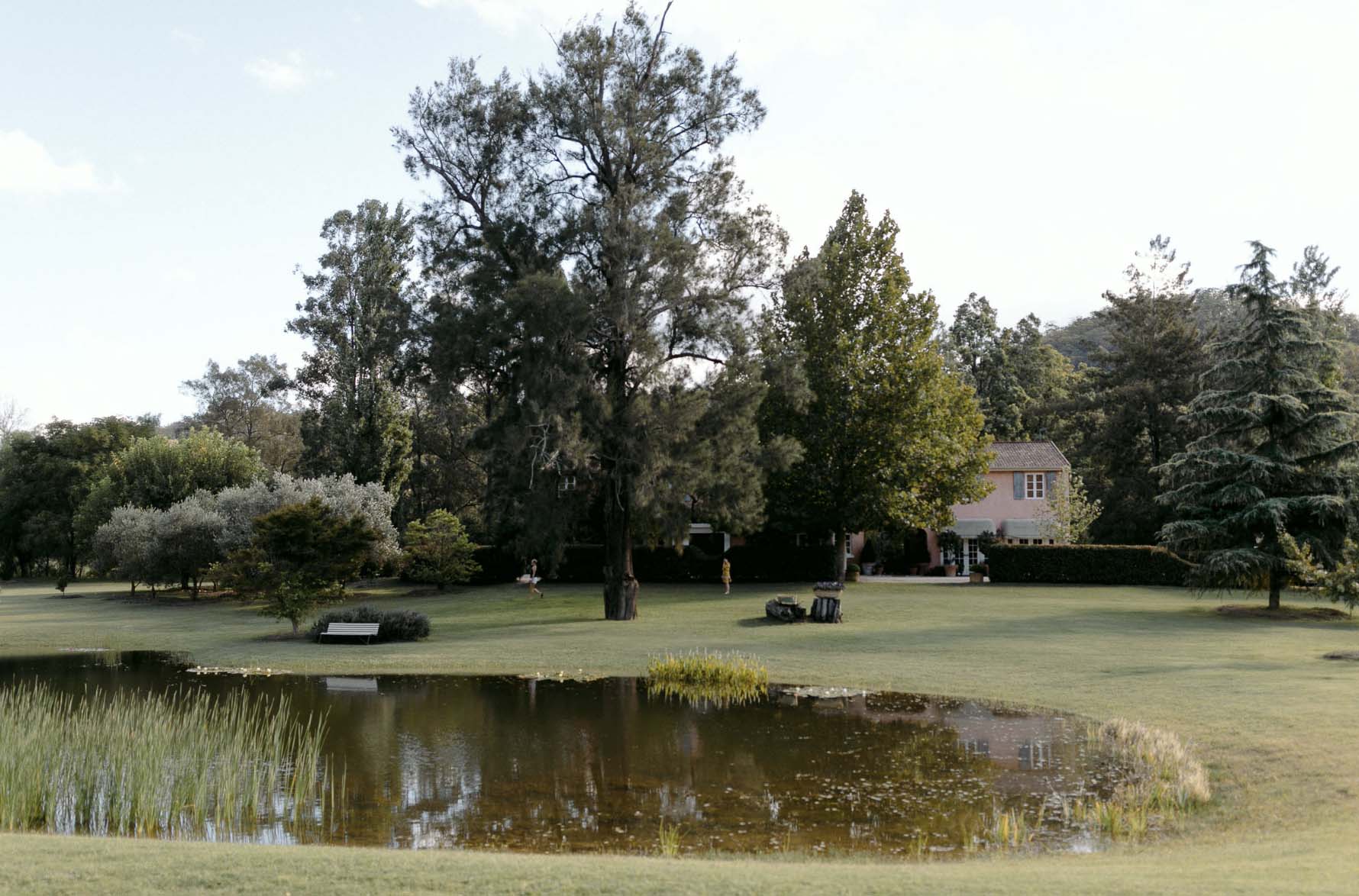 Scenic view of a pond reflecting the sky, surrounded by trees and grassy fields, with a glimpse of a building in the distance.
