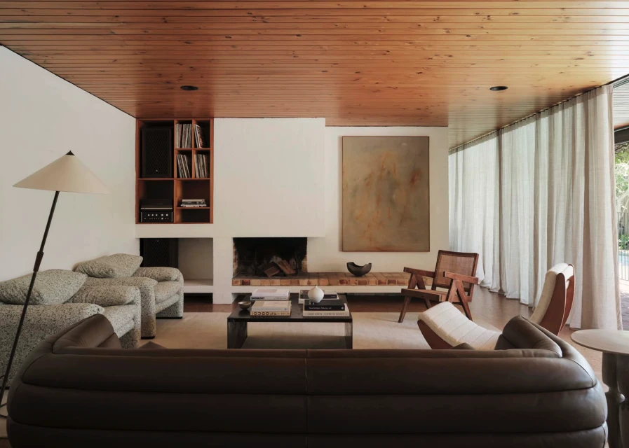 Living room interior with a brown leather sofa, fireplace, wood ceiling, large abstract artwork, and natural light from large windows.
