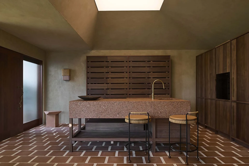 Modern kitchen interior with a terrazzo island, wooden cabinets, brick flooring, brass faucet, and natural light from a skylight.