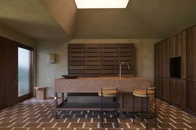 Modern kitchen interior with a terrazzo island, wooden cabinets, brick flooring, brass faucet, and natural light from a skylight.