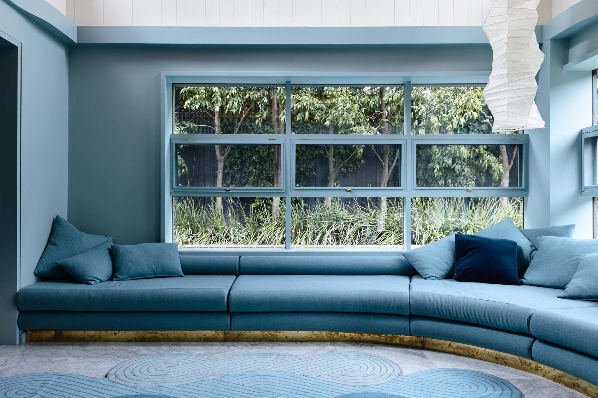Interior view of a blue-toned living room with a large multi-pane window overlooking a green garden, featuring a long, cushioned bench seat.