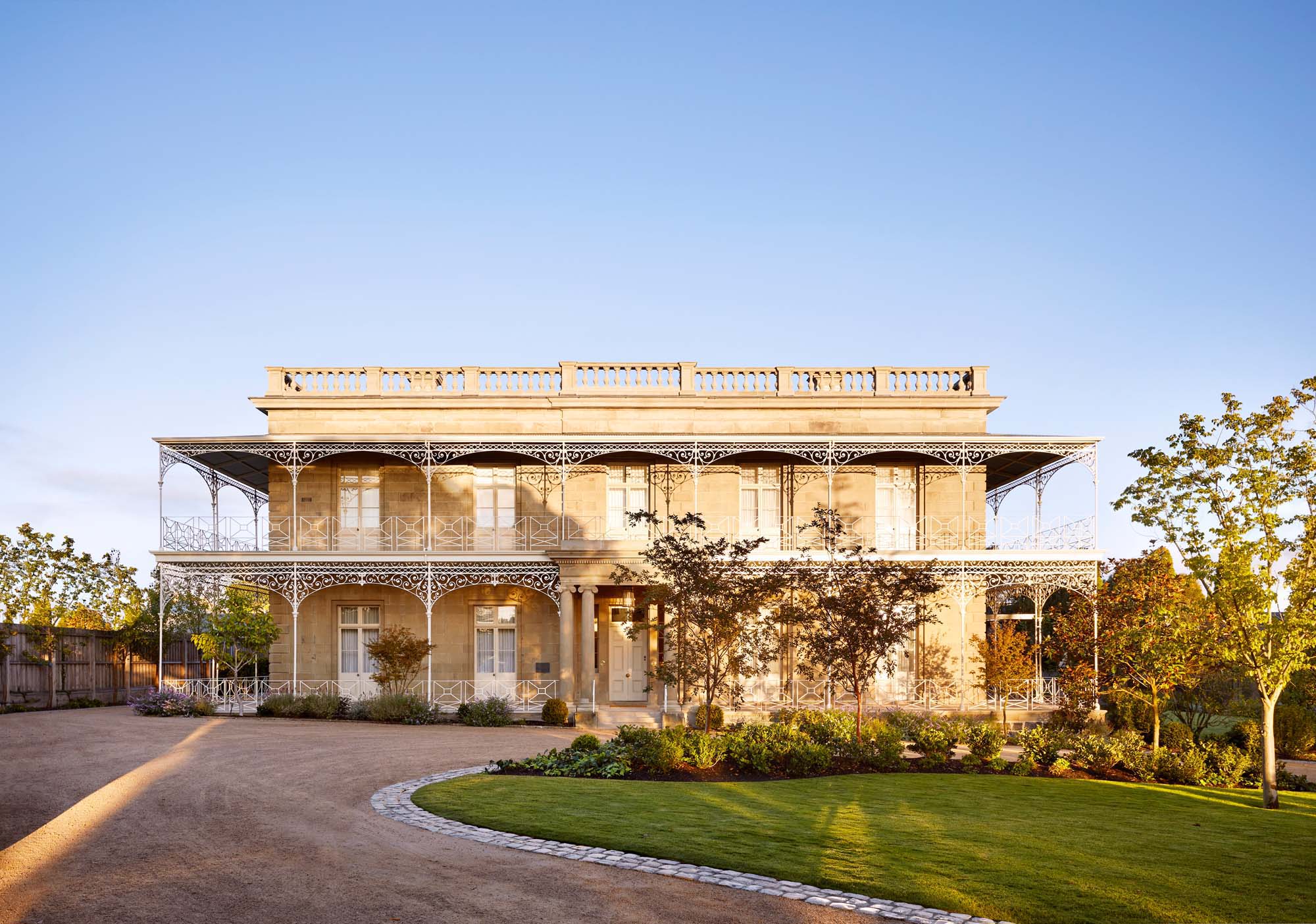Exterior view of a large, two-story beige stone house with ironwork balconies, manicured lawn, gravel driveway, and clear blue sky.