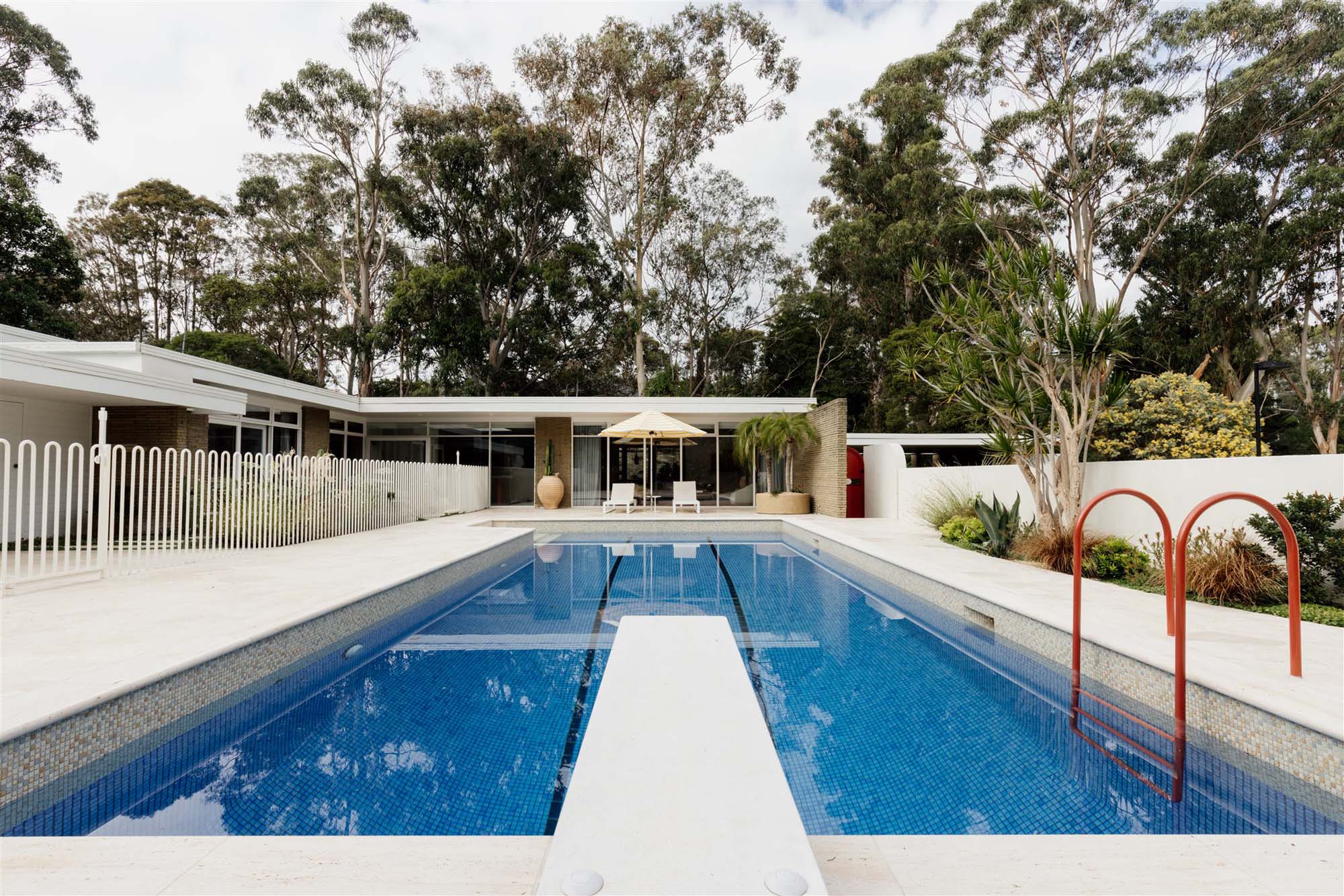 Rectangular blue swimming pool with a white diving board, red handrails, and a modern single-story house surrounded by tall trees.