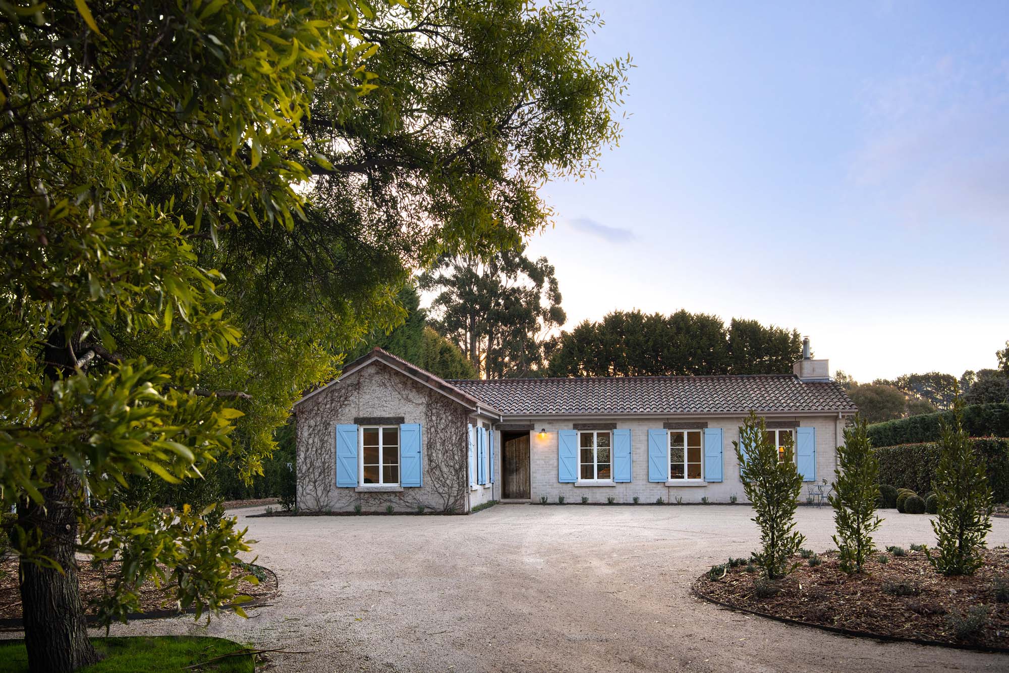 Exterior of a one-story home with a light-grey stone facade, baby-blue shutters, a gravel driveway, and green foliage surrounding the building.