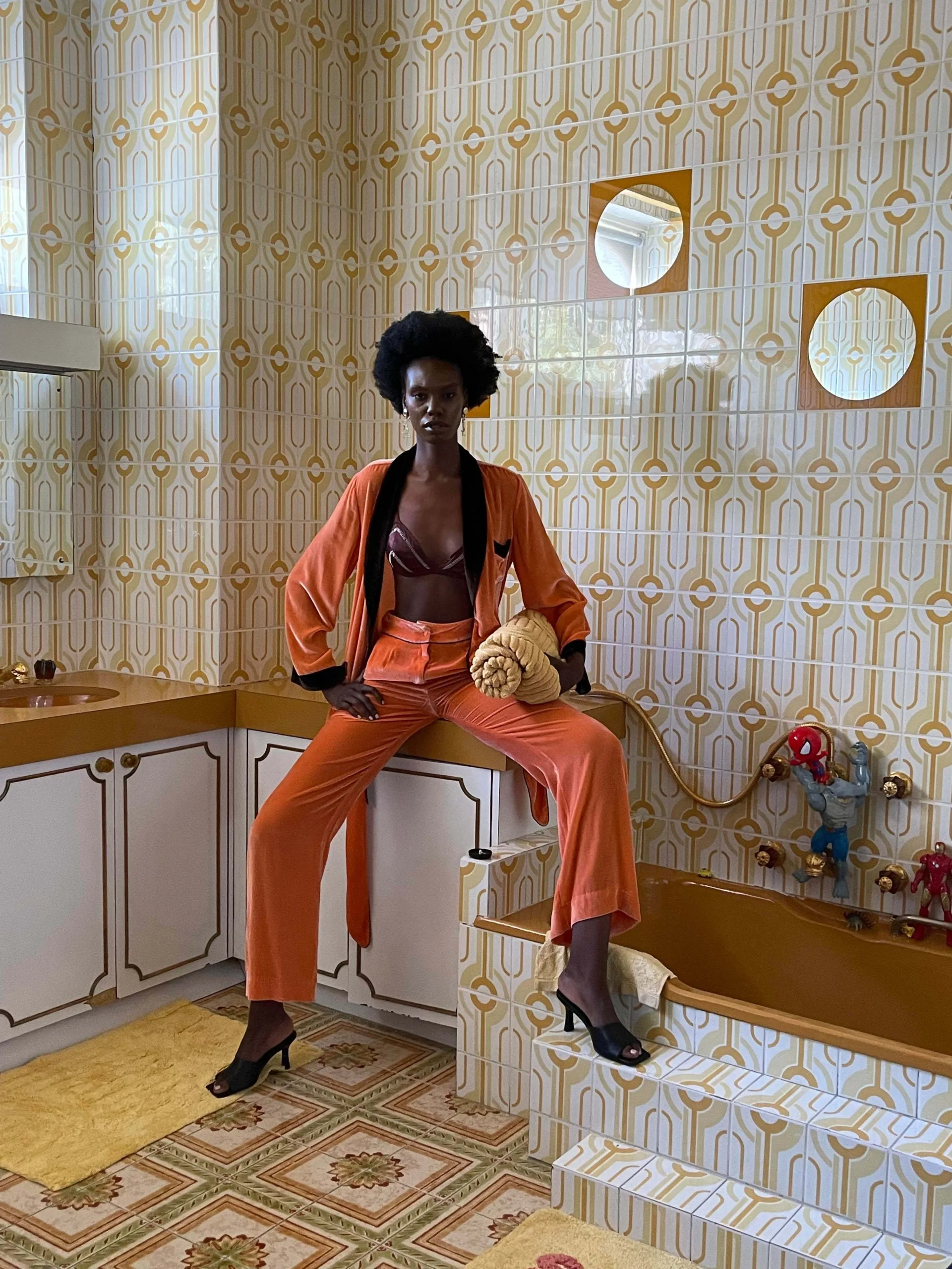An adult woman with dark skin and an afro sits on the edge of an orange bathtub, in a matching orange velvet suit, in a retro bathroom.