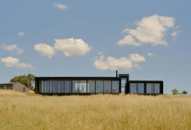 Modern black rectangular glass pavilion house set in a field of tall dry golden grass under a blue sky with scattered white clouds.