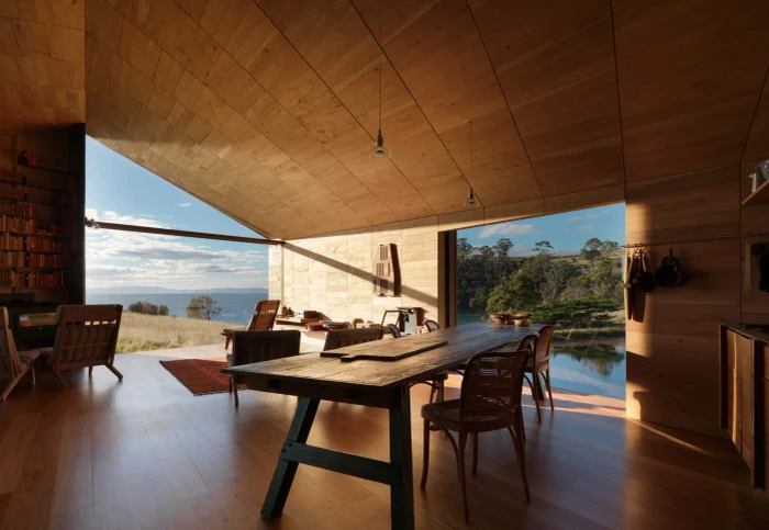 Modern wooden interior with a rustic dining table and expansive open-air windows overlooking a sunny coastal landscape.