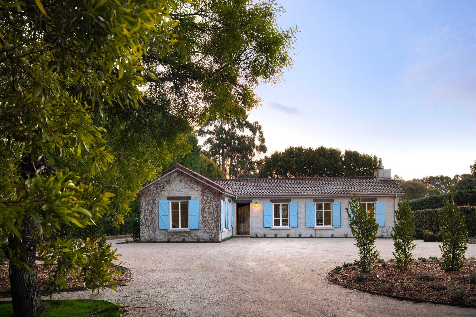 Single-story stone cottage featuring light blue window shutters, a gabled tiled roof, and an open gravel driveway.