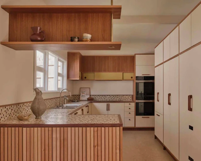 Mid-century style kitchen interior with wooden slatted counter, mosaic tile countertops, cream cabinets, and open timber shelving.