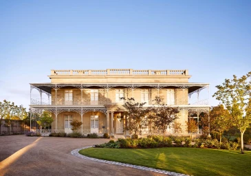 Historic two-story sandstone estate featuring ornate white iron lacework balconies and a manicured lawn under a clear sky.