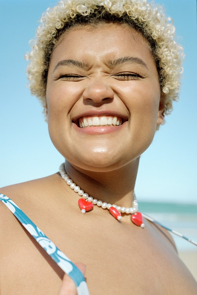 Close-up portrait of a smiling woman with blonde curly hair, wearing a blue floral top and red heart necklace, on a sunny beach.