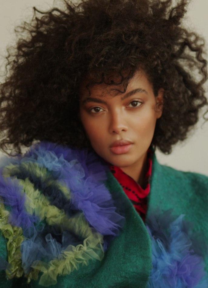 Portrait of a stylish Black woman with curly hair wearing a teal coat with blue and green ruffles, posing in a brightly lit photo studio.
